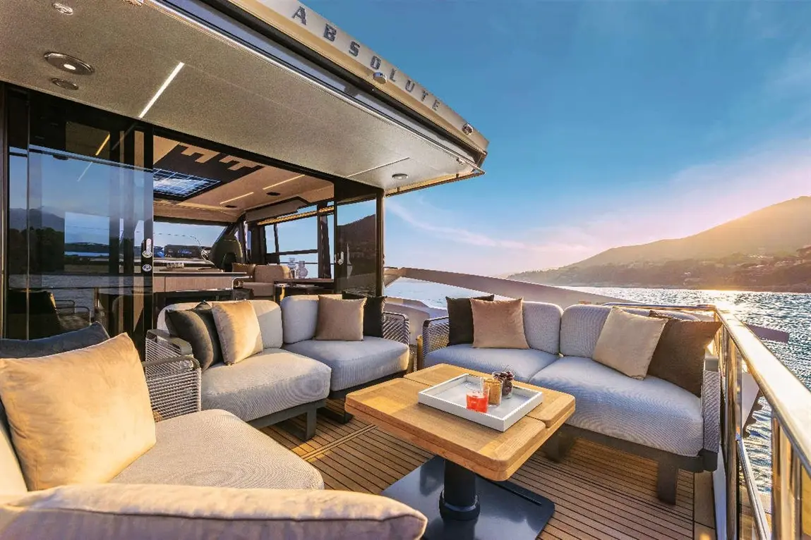 Luxury yacht lounge area with plush seating, wooden table, and drinks; scenic view of water and mountains at sunset.